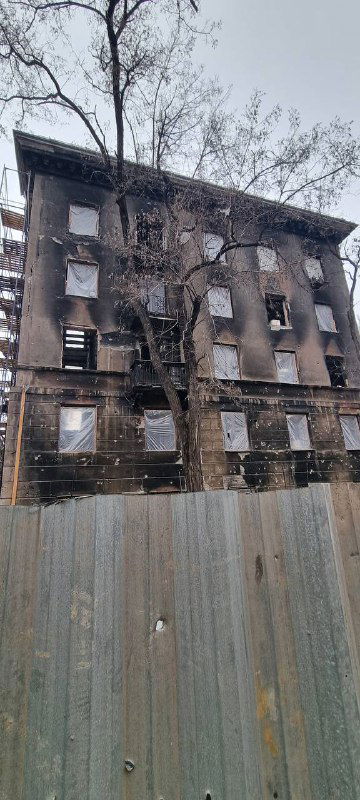Обветшалый многоквартирный дом с выбитыми окнами и следами разрушений; вид сбоку через временное ограждение и опавшие ветки деревьев.