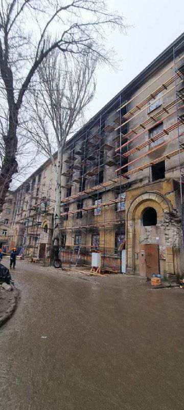 Строгое изображение фасада многоэтажного здания с частичными разрушениями и ограждением вдоль улицы — городской пейзаж Мариуполя в зоне работ.