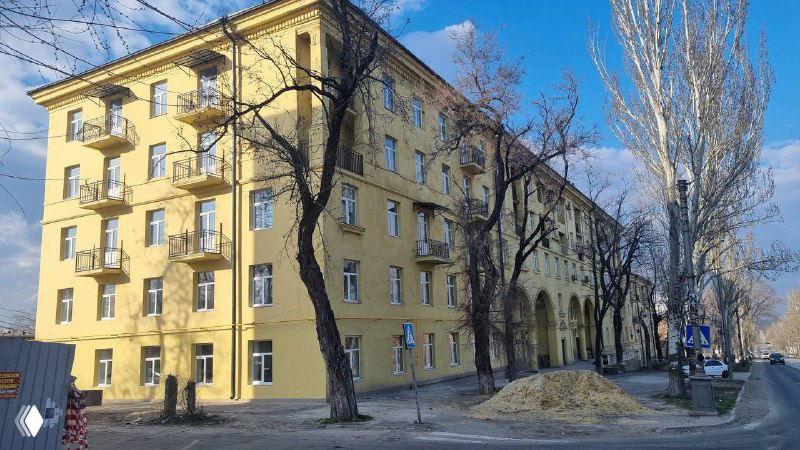 Отреставрированный фасад жилого дома, окрашенный в светлый тон; вид улицы с деревьями и восстановленным архитектурным обликом здания.