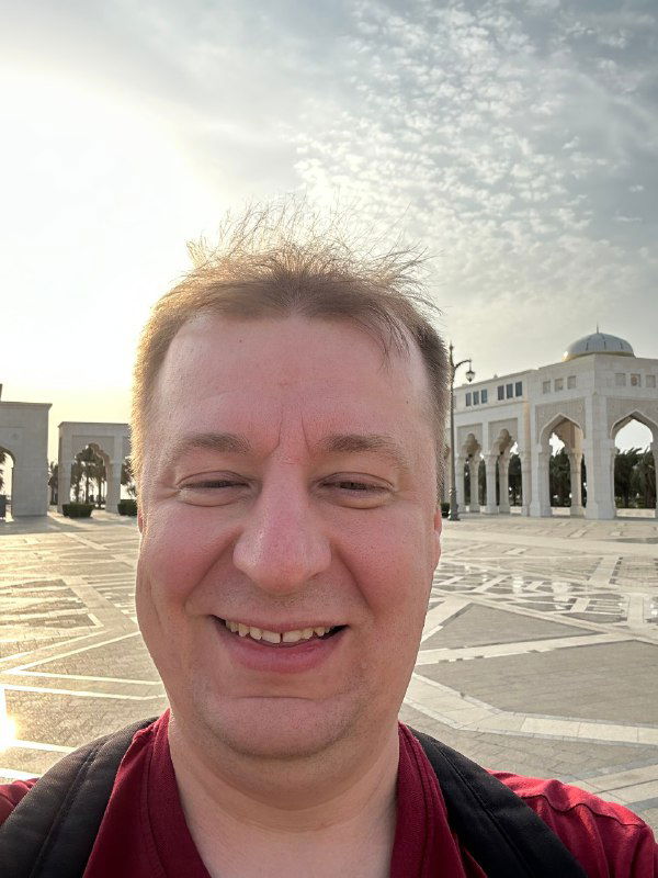 Портрет улыбающегося мужчины среднего возраста на открытой площади с архитектурными арками и светлым небом на заднем плане, дневной кадр