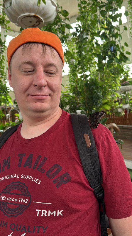 Селфи мужчины в оранжевой шапке и с рюкзаком на плече в интерьере с зелёными растениями, видны детали одежды и расслабленная улыбка