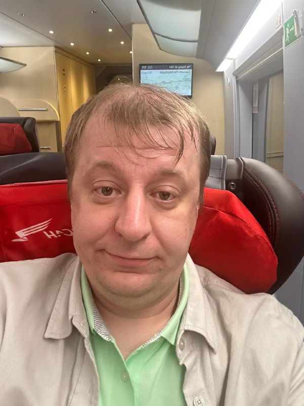 Крупный план человека в вагоне поезда: лицо в кадре, красные спинки кресел и интерьер современного поезда, дневное освещение через окно в фоне.