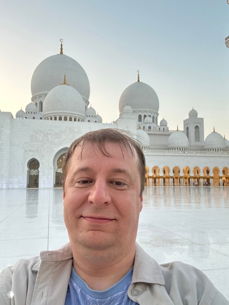 Селфи мужчины на открытой площади перед белыми куполами и фасадом мечети; мягкий вечерний свет и отражение на мраморной плитке вокруг.