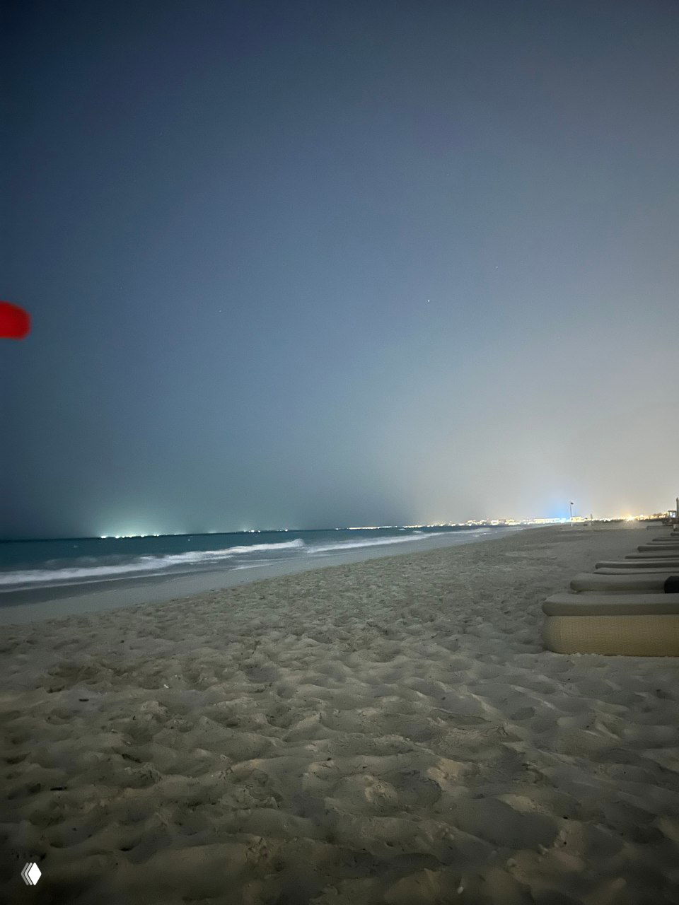 Ночной пляж с ровным песком и легким прибоем; вдали видны огни города по линии горизонта и приглушённая морская гладь.