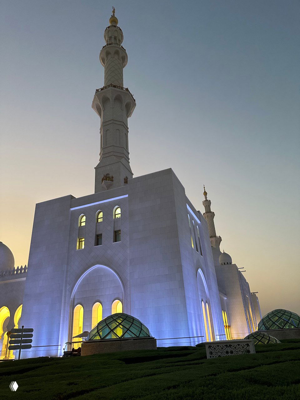Вид снаружи на мечеть в сумерках: высокий минарет, освещённый фасад и арочные окна, подчёркнутые голубой подсветкой и тёплым вечерним небом.