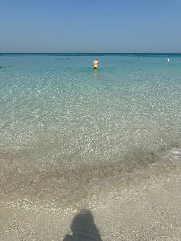 Чистая бирюзовая вода и мелкий песок: человек стоит в море на фоне спокойного пляжа в тёплом регионе, утреннее или дневное освещение.