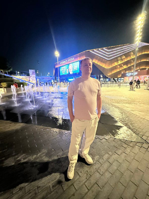 Мужчина стоит вечером перед подсвеченной Etihad Arena: фасад арены, фонтаны и ночные огни создают атмосферу перед концертом.