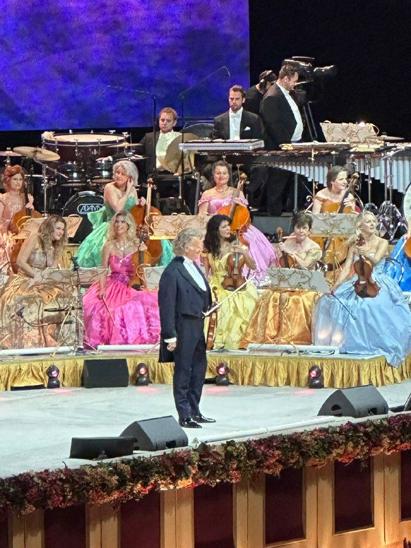 Сцена концерта Andre Rieu: дирижёр в центре перед большим оркестром и разноцветными костюмами музыкантов на помпезной сцене.