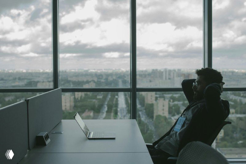 Человек в офисе у окна с ноутбуком на переднем плане и городским пейзажем за стеклом, символ выбора и перспектив на рынке работы