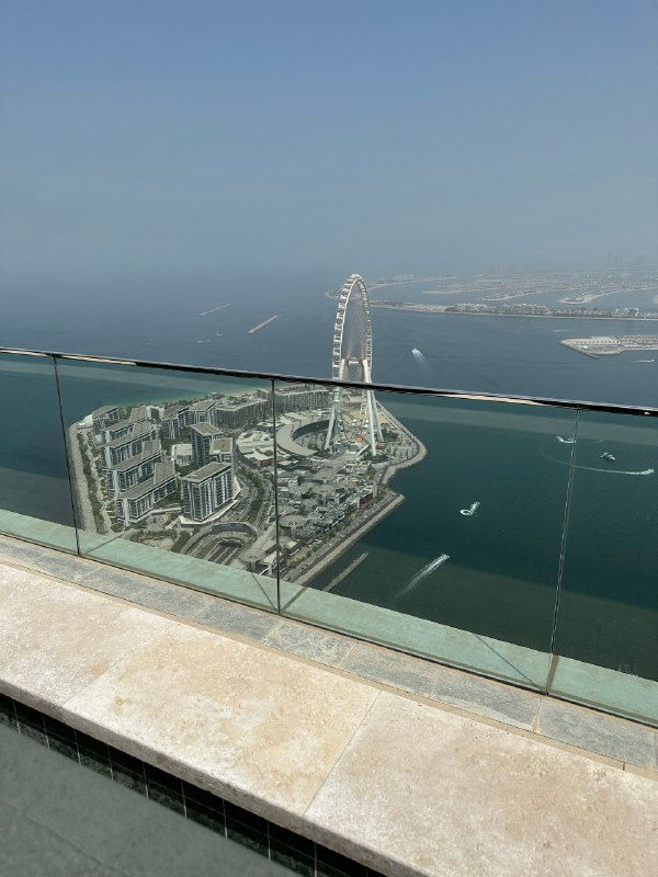 Съёмка с высоты показывает стеклянный парапет балкона, морской берег и городскую застройку; вдалеке виден арочный небоскрёб и блеск моря.