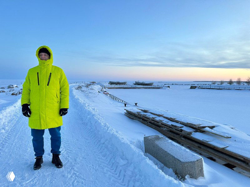Человек в ярком неоново-зелёном пуховике стоит на заснеженной дороге; вокруг простёрлись снежные поля, иней и холодный закатный пейзаж.