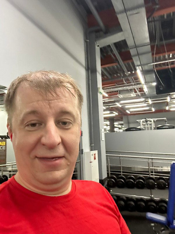 Селфи автора в спортзале в красной футболке на фоне тренажёров и потолочных светильников — неформальный портрет коуча.