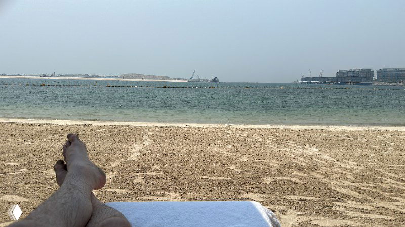 Пляжный кадр с вытянутыми ногами на шезлонге, песчаным берегом и спокойным морем; кадр передаёт атмосферу отдыха и расслабления на побережье.