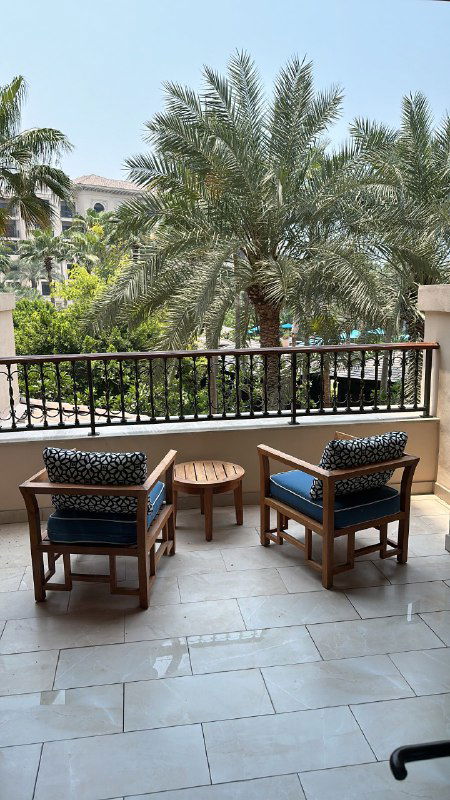 Балкон апартаментов с плетёной мебелью, столиком и видом на пальмы и зелёную листву; тёплая атмосфера отдыха в тропическом окружении.