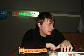 Фотография молодого человека в тёмной рубашке, боковой ракурс за столом; интерьер бара или кафе на заднем плане, личный архив, неинформативные детали.