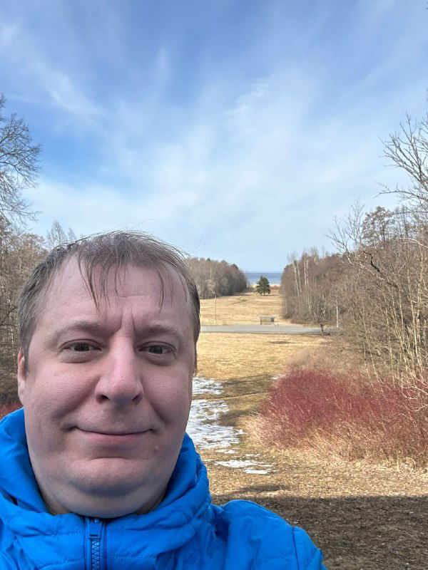 Селфи мужчины в синей куртке на фоне открытого холма и просеки с редкими кустами, видна весенняя природа и остатки снега под небом.