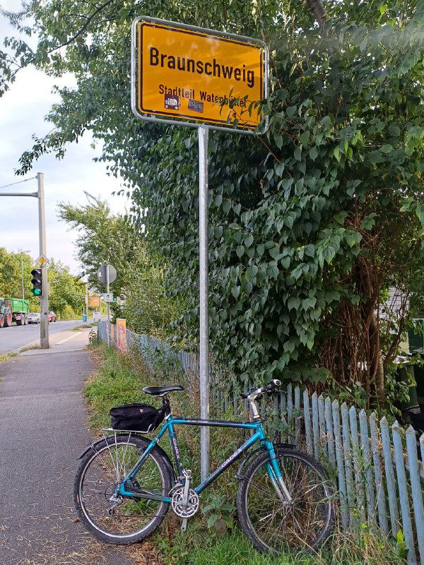 Голубой байк припаркован у дорожного знака «Braunschweig» на фоне зелёных кустов и деревянного забора, сельская сцена и тротуар.