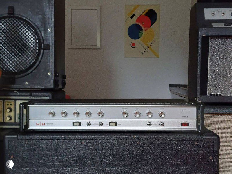 Фронтальная панель винтажного усилителя HH IC100S на стэке с видимыми регуляторами и металлическим корпусом в комнате с плакатом.