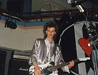 Музыкант с электрогитарой на сцене во время выступления, видна часть сцены, усилители и плакат на стене, атмосферный снимок живого концерта.