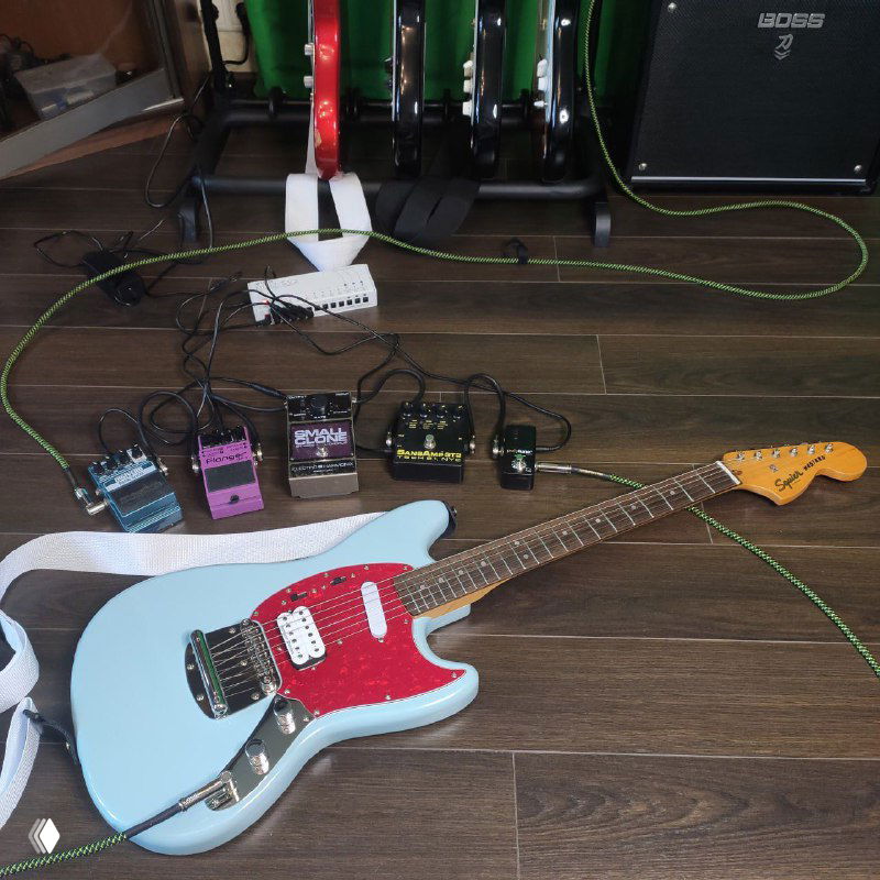 Голубая Squier Mustang Classic Vibe на полу рядом с эффект-педалями и проводами; видна задняя часть и пикгард.