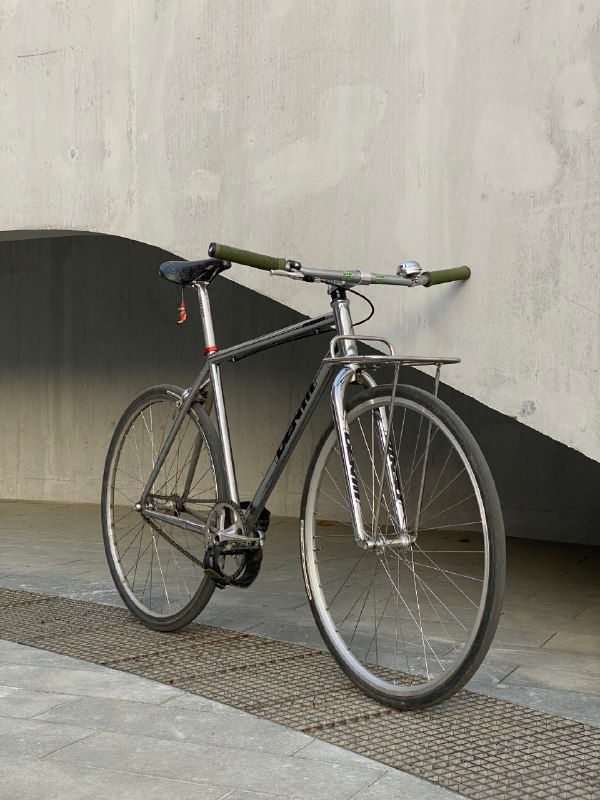 Боковой ракурс серебристого фикса Identiti на урбанистическом фоне: видна рама, руль с прямой планкой и тонкие колёса, минималистичный стиль.