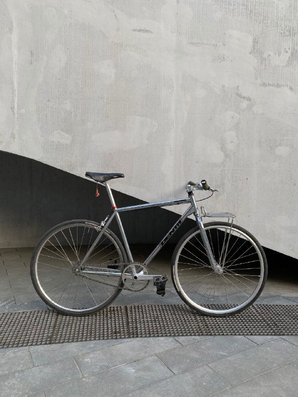 Полный кадр велосипеда Identiti у бетонной стены: тонкая рама, односкоростная трансмиссия и классическая посадка, городской минимализм.