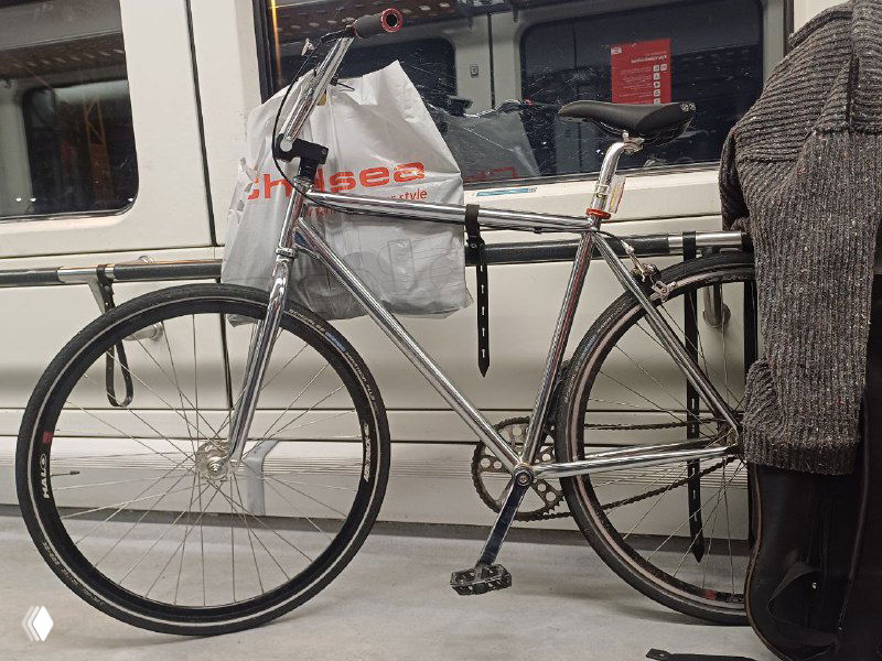 Фикс-байк Identiti, пристёгнутый в вагоне или на платформе: рама и колёса в состоянии после поездки, пакет на багажнике и повседневный городской антураж.