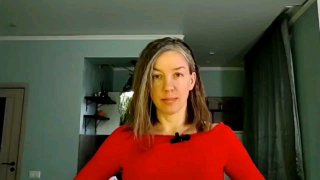 Женщина в красном пуловере стоит перед камерой в домашней комнате, видны полки, занавески и мягкое освещение на заднем плане.