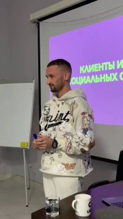 Мужчина на мероприятии выступает у экрана с фиолетовым слайдом о клиентах и соцсетях; рядом белая доска и кружка.