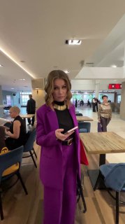 Женщина в фиолетовом костюме в холле мероприятия держит телефон; на заднем плане посетители, столы и зона для общения участников.