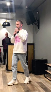 Спикер в светлом свитшоте стоит с микрофоном на сцене офлайн‑мероприятия в Тюмени; видна сцена, техническое оборудование и часть аудитории в зале.