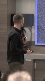 Мужчина с микрофоном стоит боком у экрана презентации в зале — кадр с выступления, символ действия и энергии.