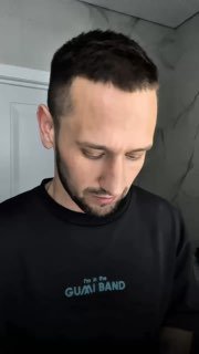 Человек в тёмной футболке, смотрящий вниз в ванной комнате; на фоне светлая плитка и дверь, нейтральная мимика, портретный кадр сверху, мягкое освещение