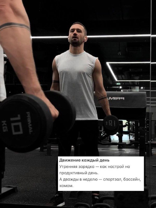 Мужчина в спортзале поднимает гантели на фоне зеркал и металлической арматуры; снимок иллюстрирует привычку движения и поддержания физической формы.