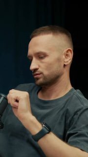 Крупный план ведущего в студии подкаста: мужчина с короткой стрижкой держит руку у микрофона на тёмном фоне.