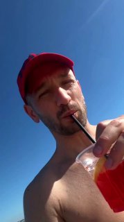 Автор канала в красной кепке и без футболки на фоне чистого синего неба, пьёт яркий напиток через трубочку.