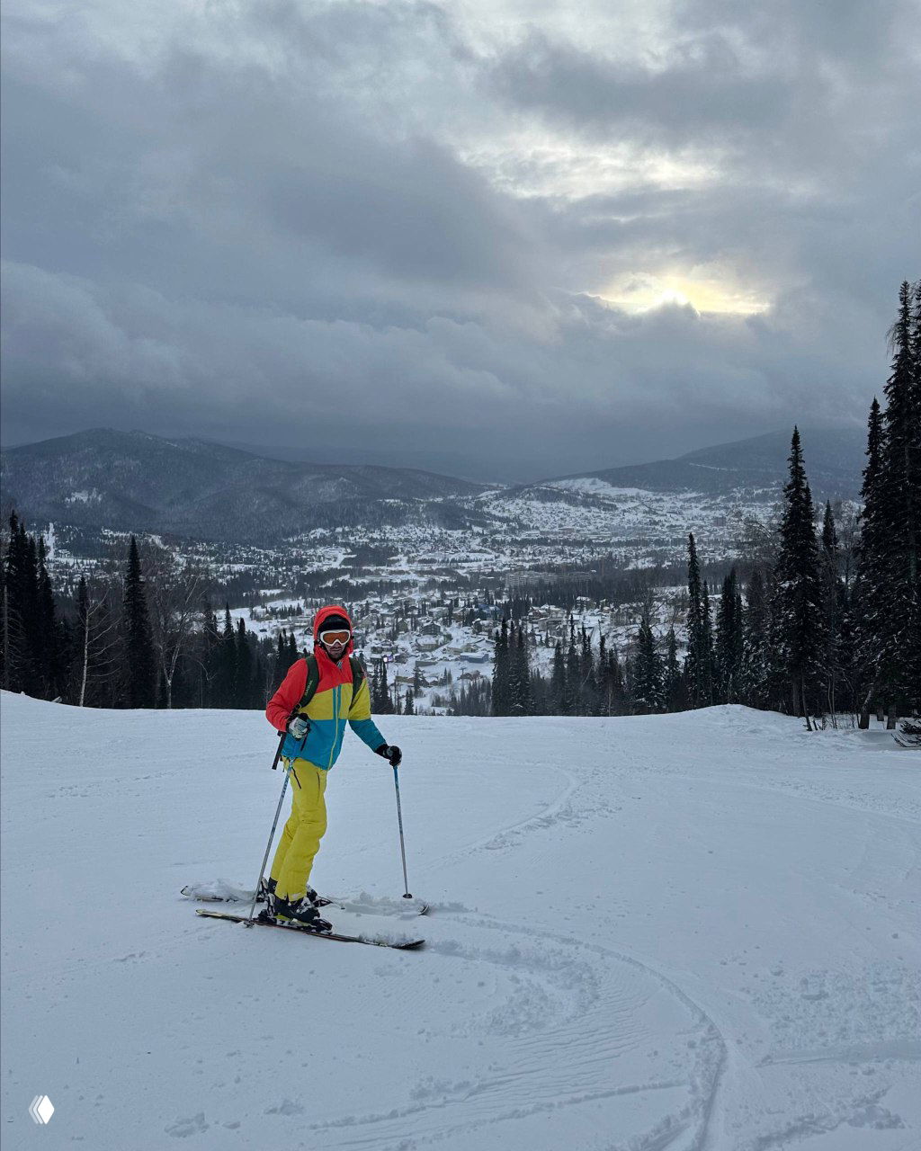 Человек на лыжном склоне в яркой экипировке на фоне заснеженных гор и пасмурного неба; кадр иллюстрирует увлечение горнолыжным спортом.