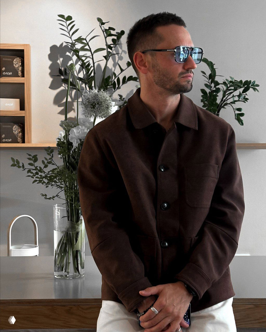 Портрет в интерьере с растениями и вазой цветов, мужчина в коричневой куртке смотрит в сторону; атмосферный кадр стильного повседневного образа.