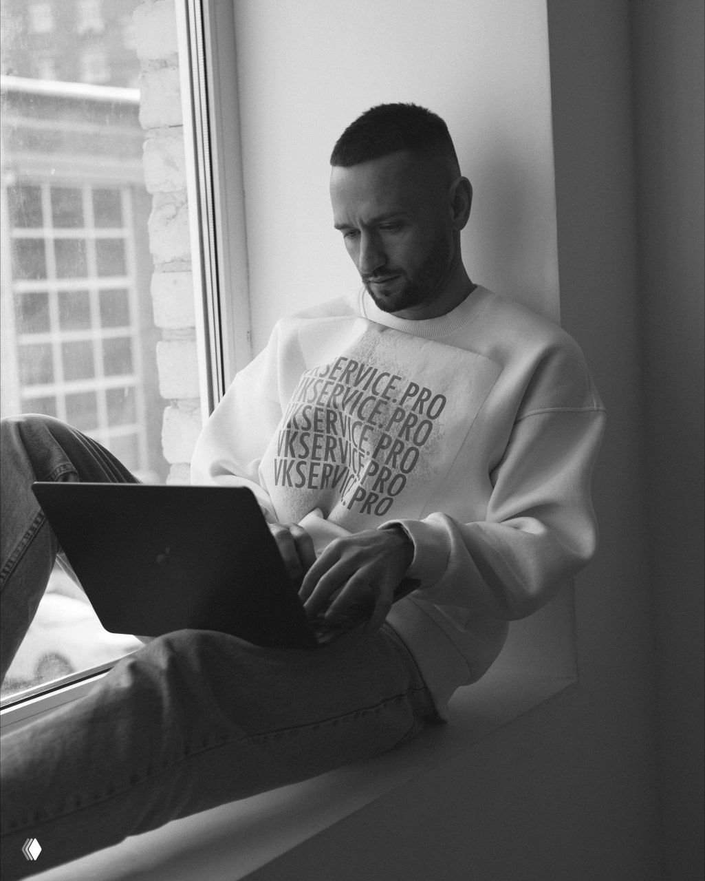 Чёрно‑белый кадр: мужчина работает за ноутбуком на подоконнике; спокойная рабочая сцена отражает фокус на IT и удалённой работе.