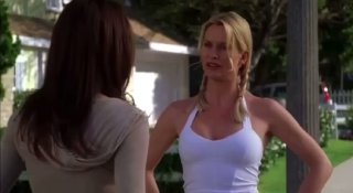 Кадр из сериала «Отчаянные домохозяйки»: женщина в белой майке и косичках разговаривает на улице перед домом, эмоциональное столкновение между персонажами.