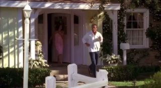 Кадр из сериала «Отчаянные домохозяйки»: персонаж выходит из дома на веранду, видны фасад дома, сад и белый забор, типичная субурбанная сцена.