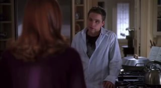 Сцена на кухне: женщина смотрит в сторону молодого мужчины у плиты, напряжённая бытовая сцена, разговор в семейной обстановке.