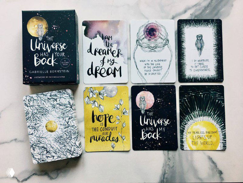 Набор небольших вдохновляющих карточных оракулов, разложенных на мраморном фоне: карты с мотивационными надписями и иллюстрациями в пастельных тонах.