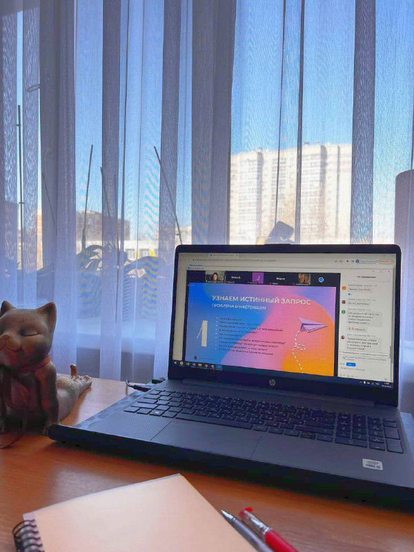 Ноутбук на столе с тем же слайдом тренинга, рядом фигурка кота, блокнот и ручки; вид на окно и городской пейзаж за тонкой шторой