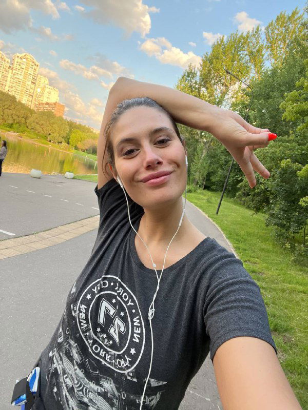 Селфи той же женщины в парке с поднятой рукой; наушники, тёмная футболка с принтом, зелёные деревья и высотки на заднем плане.