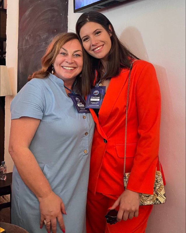 Автор с другой участницей конференции в тёплой дружественной обстановке, обе улыбаются, на шее видны бейджи, интерьер зала на фоне.