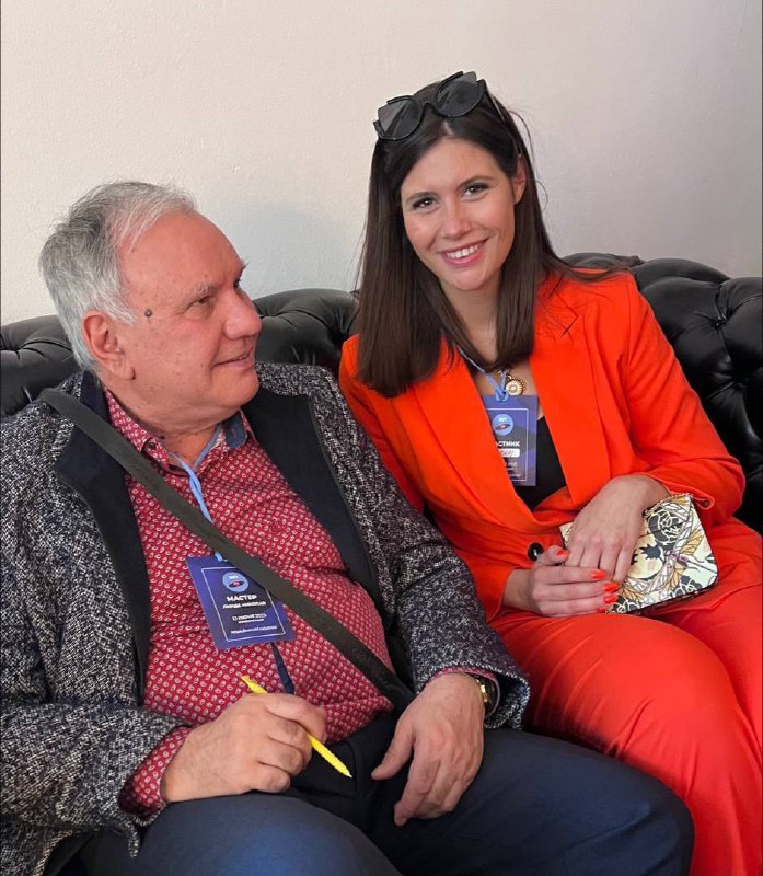 Автор фотографируется рядом с пожилым мужчиной в роли лектора или участника: они сидят на диване, у обоих видны бейджи и ручка для заметок.