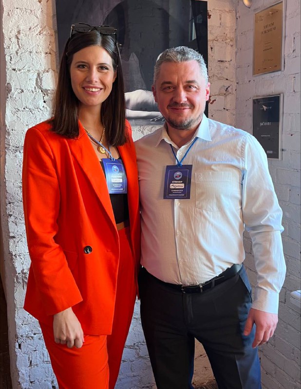 Автор стоит рядом с мужчиной в деловой одежде в интерьере конференц-пространства, оба улыбаются, на них видны бейджи участника.