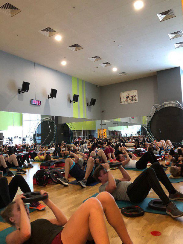 Групповая тренировка в зале: участники лежат на ковриках и делают упражнения пресса, в кадре зеркала и спортивный реквизит.