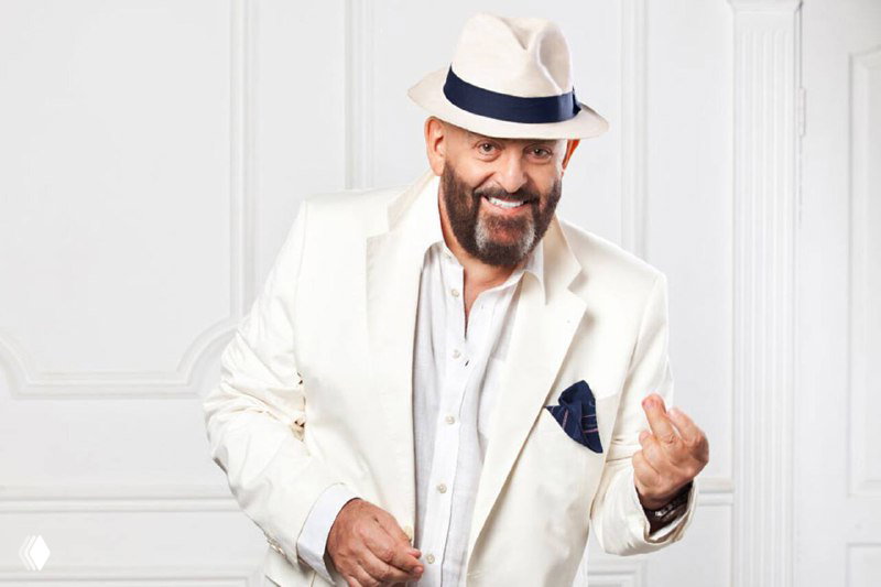 Портретный кадр улыбающегося мужчины-исполнителя в белом костюме и шляпе: студийная фотография в праздничной стилистике.